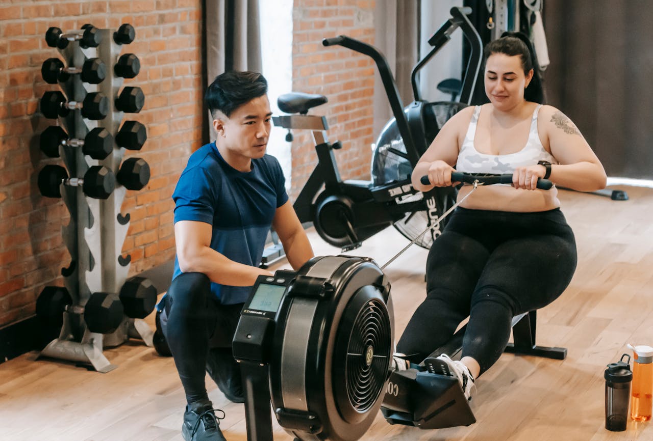 about-us Positive young plus size female in leggings and crop top doing abs exercise on rowing machine during intense workout in modern gym with fit Asian male trainer